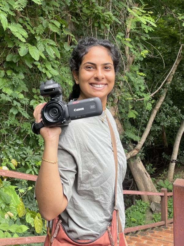 Norah Rami, pictured holding a camera in the foreground. Behind her, trees from a forrest and a flight of stairs.