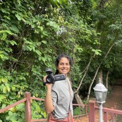 Norah Rami, pictured holding a camera in the foreground. Behind her, trees from a forrest and a flight of stairs.